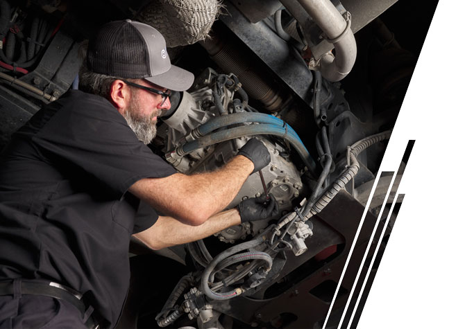 A man dressed in black with a black and gray baseball hat and black gloves uses a tool to work on a transmission under a vehicle.